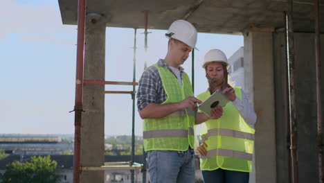 Engineers-builders-man-and-woman-standing-on-the-roof-of-the-building-with-a-tablet-computer-discussing-in-white-helmets-and-shirts.
